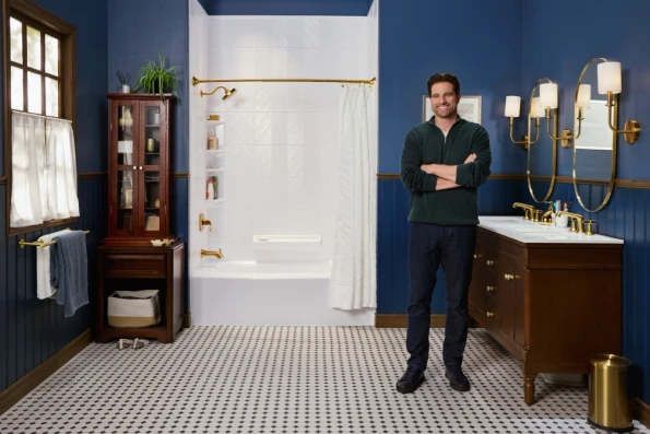 A smiling man stands with his arms crossed in a newly remodeled bathroom. The room features dark blue walls, a white and black tiled floor, and gold fixtures. The man is standing in front of a white bathtub and shower surround, and next to a wooden vanity with a white countertop and a mirror with gold light fixtures.